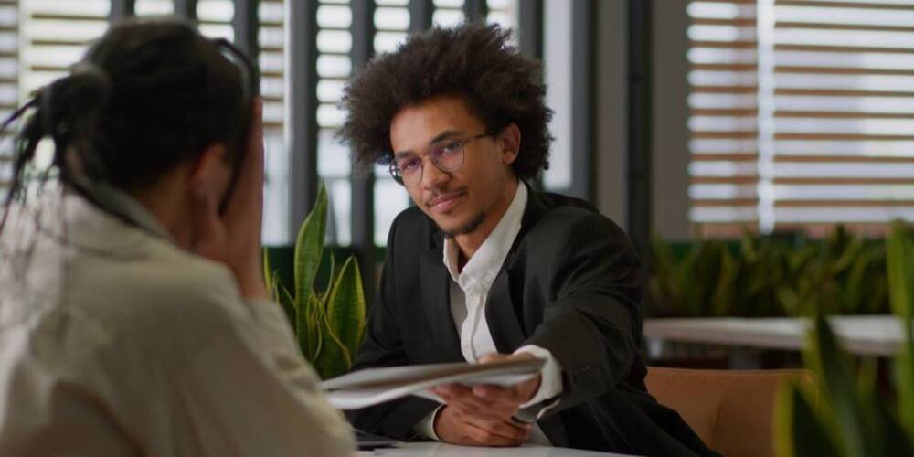 Man with head in hands opposite man handing him paper